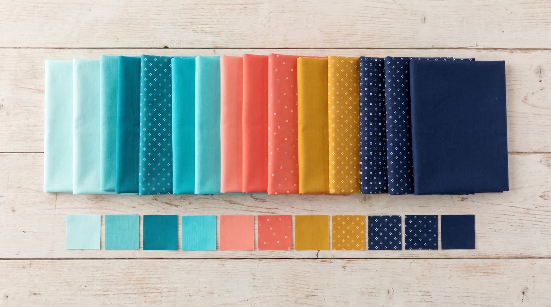 Folded quilting fabrics arranged in a gradient from pale aqua through coral and mustard to navy, with small cut squares below